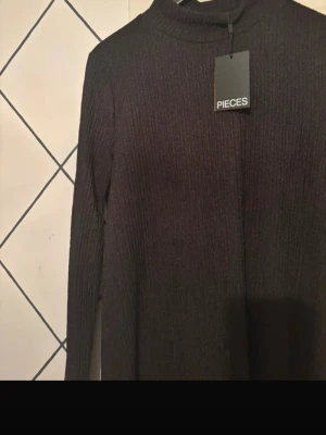 Svart ribbad långklänning Pieces - Snygg svart långklänning från Pieces med ribbad struktur och lång ärm. Klänningen har en hög hals och är i ett mjukt, stretchigt material som sitter bekvämt. Perfekt för dig som gillar stilrena och enkla plagg.