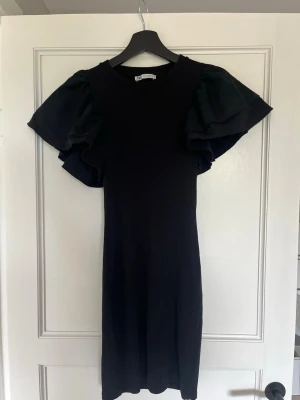 Svart klänning med volangärmar Zara - Svart klänning från Zara med stora volangärmar och rund halsringning. Klänningen har en rak passform och är tillverkad i ett ribbat material som ger en snygg struktur. Perfekt för dig som gillar stilrena plagg med en twist.