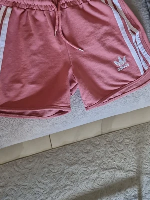 Rosa Adidas shorts med vita ränder - Säljer ett par snygga rosa Adidas shorts med vita ränder på sidorna och klassisk Adidas-logga broderad framtill. Shortsen har elastisk midja med snörning och är tillverkade i mjuk bomull, perfekta för en sportig look. Deb är mindre I storlek 