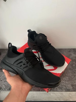 Svarta Nike Presto sneakers - Snygga helsvarta Nike Presto sneakers med låg profil och sportig design. Skorna har en ovandel i mesh och syntet, rund tå och klassisk snörning. Swoosh-logga på sidan och tå. Perfekta för dig som gillar stilrena och bekväma sneakers.