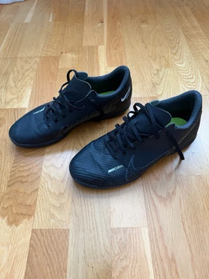 Svarta Nike Mercurial fotbollsskor - Säljer ett par svarta Nike Mercurial fotbollsskor med snörning. Skorna har en stilren design med diskreta vita detaljer och Nike-logga på sidan. Ovandelen är tillverkad i syntetmaterial för bra bollkontroll och passform.
