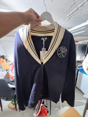 Marinblå kofta med tennismärke - Stilren marinblå kofta med dragkedja och krage i vitt med svarta ränder. På bröstet sitter ett broderat tennismärke med två korsade racketar. Perfekt för en sportig och preppy look. Finns att hämta i Gröndal. 