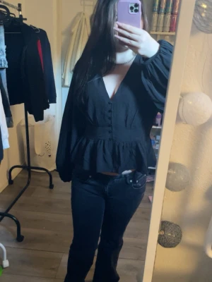 Svart blus med volang och knappar - Snygg svart blus med v-ringning, dekorativa knappar framtill och volang nedtill från lager 157. Blusen har långa ärmar och lätt puffig axel för en trendig look. Perfekt att styla med jeans eller kjol för en chic outfit. Använt en gång🤍 pris kan självklart diskuteras💕