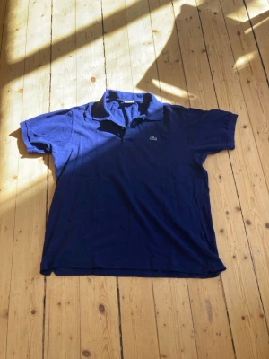 Marinblå pikétröja från Lacoste - Snygg marinblå pikétröja från Lacoste med klassisk krage och korta ärmar. Tröjan har det ikoniska krokodilmärket broderat på bröstet och är tillverkad i bomull för en bekväm känsla.