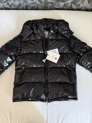 Moncler Maya  - Snygg svart dunjacka från Moncler med glansig finish, huva och dragkedja framtill. Jackan har två framfickor med dragkedja och en ficka med Moncler-logga på ärmen. Perfekt för kalla dagar och riktigt trendig look. Detta är en moncler Maya som aldrig är använd och passar perfekt till vintern, jackan är i storlek 1/S. NFC finns även.