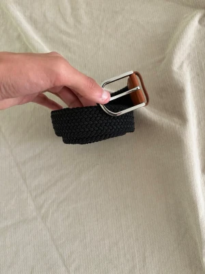 Svart flätat bälte med silverfärgat spänne - Snyggt svart flätat bälte med silverfärgat metallspänne och bruna detaljer i skinnimitation. Bältet är flexibelt och passar till många olika stilar. Längd 120 cm enligt märkning.