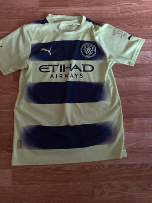 Manchester City matchtröja  - Snygg Manchester City matchtröja från Puma i grön med breda mörkblå ränder. Tröjan har klubbmärke och Puma-logga på bröstet samt Etihad Airways-tryck på framsidan. Tillverkad i ett lätt och ventilerande material, perfekt för fotboll eller träning.