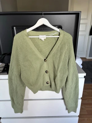 Grön ribbstickad kofta med knappar - Snygg ljusgrön ribbstickad kofta med v-ringning och tre stora bruna knappar framtill. Koftan har lång ärm och en croppad passform som ger en trendig look. Den är köpt second hand men märket är Monki. I storlek XS.