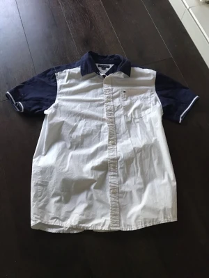 Vit och marinblå kortärmad skjorta från tommy hilfiger - Kortärmad skjorta i vitt med marinblå ärmar och krage från tommy hilfiger. Skjortan har knappar framtill och tryck på ryggen Tillverkad i bomull och har en klassisk passform.