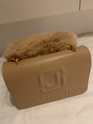 Beige handväska med fuskpälsdetalj - Snygg beige handväska med mjukt handtag i fuskpäls och guldfärgade metalldetaljer. Väskan har en stilren, boxig form och präglad logga på framsidan. Perfekt accessoar för dig som vill sticka ut med en trendig och unik look.