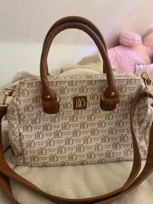 handväska  - Snygg beige handväska från Lycke Oslo med bruna detaljer och dubbla handtag. Väskan har ett all-over logomönster, guldfärgade metalldetaljer och avtagbar axelrem. Perfekt för dig som gillar stilrena accessoarer med en touch av lyx.