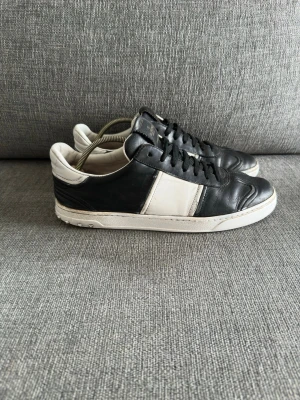 Valentino garavani flycrew sneckers läder svart/vit. Obs ej orginal innesula -   Valentino Garavani  Storlek: 42 Färg: Svart / Vit  Material: Läder  Sulan: Det finns ett tydligt släpp/spricka i det yttre gummilagret under hälen på båda skorna (se bild 4).  Hälparti: Visst slitage/skav finns på det vita lädret på hälens insida och ovankant, samt mindre skavmärken på den lädret. Sulkräm har andvänts och i stor del dåligt skick. Lädret har också blivit avfärgat från vit till en mörkare nyans. Undersulan har också flera hål fram och bak. Majoriteten av nitarna är inte orginal.