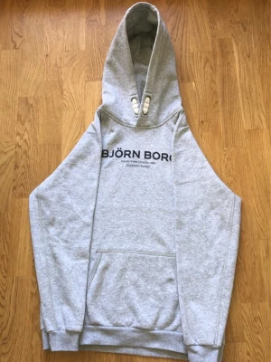 Grå hoodie från Björn Borg - Snygg grå hoodie från Björn Borg med svart tryck på bröstet där det står 'BJÖRN BORG Autumn/Winter Collection 1984 Stockholm Sweden'. Hoodien har stor huva, magficka och ribbade muddar. Perfekt för chill dagar eller när du vill ha en clean och enkel stil.