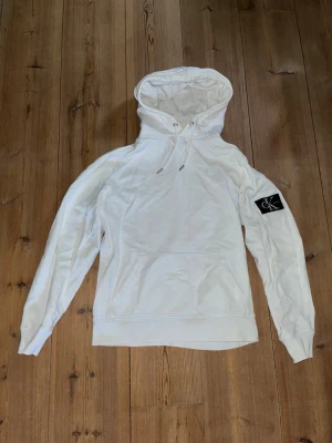 Vit hoodie från Calvin Klein  - Snygg vit hoodie från Calvin Klein med huva. Stor CK-logga på vänster ärm ger en clean och modern look. Hoodien har en clean magficka och ribbade muddar. Perfekt för dig som gillar stilrena plagg med en clean look! Perfekt för hösten!  Aldrig använd endast testat och liggandes i garderob! Nypris:1299kr.     Mitt pris:699kr