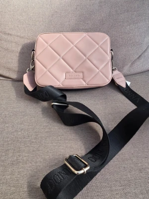 Rosa quiltad axelväska Don Donna - Snygg ljusrosa axelväska från Don Donna i quiltad design. Väskan har en bred, justerbar svart axelrem med logga och silverfärgade detaljer. Tillverkad i ett slätt, konstlädermaterial och har en stilren, rektangulär form.  Mått 21x16x6