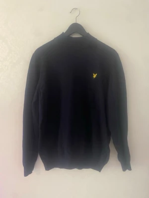 Lyle & Scott mörkblå tröja, storlek XL - En stilren mörkblå tröja med rund hals och lång ärm. På bröstet finns en liten gul broderad logga som ger en snygg detalj. Tröjan har ribbade muddar vid ärmslut och nederkant. Perfekt till jeans eller chinos.
