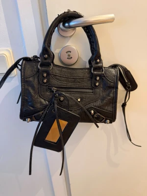 Svart city bag med nitar och spegel - Cool svart handväska i skinn med silverfärgade nitar och dragkedja framtill. Väskan har dubbla handtag, dekorativa sömmar och en avtagbar spegel. Perfekt för dig som gillar edgy detaljer och vill ha en väska som sticker ut.