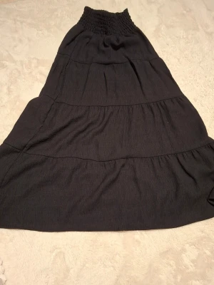 Svart långkjol med smock i midjan - En svart långkjol med smockad midja och flera volanger. Kjolen har en luftig och flowy passform, perfekt för dig som gillar en avslappnad men ändå stilren look. Passar till många olika outfits och är lätt att matcha.