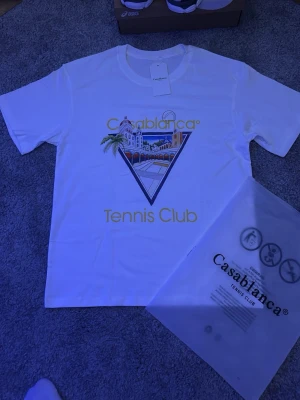Casablanca Tennis Club t-shirt vit - Vit t-shirt från Casablanca med färgglatt tryck framtill som visar en byggnad och palmer i triangelformad ram. Texten 'Casablanca Tennis Club' i gult. Den är stilren och passar perfekt till skolstarten! Fraktar inom 0-1 dagar📦🤝