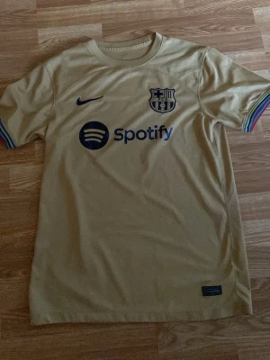 FC Barcelona matchtröja  - Snygg gul FC Barcelona matchtröja från Nike med Spotify-logga på bröstet och klubbmärke. Tröjan har korta ärmar med regnbågsfärgade detaljer vid ärmsluten. Perfekt för dig som älskar fotboll och vill visa ditt stöd för Barca.