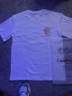 Vit Casablanca Airways t-shirt - Säljer en vit t-shirt från Casablanca med trycket 'Casablanca Airways' och en färgglad grafik med apelsin och blad både fram och bak. Klassisk passform och korta ärmar, perfekt för en clean och fräsch look.