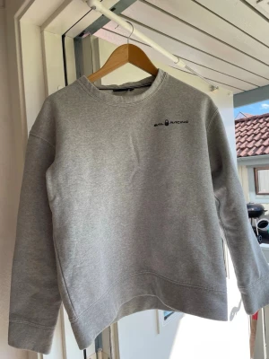 Grå sweatshirt från Sail Racing - En stilren grå sweatshirt från Sail Racing med svart logga på bröstet. Tröjan har rund halsringning, raka ärmar och är gjord i mjukt bomullsmaterial. Perfekt för en avslappnad och sportig look.