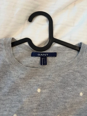 Grå stickad tröja med prickar GANT - Supersnygg grå stickad tröja från GANT med vita prickar över hela tröjan. Rund halsringning och långa ärmar. Perfekt till jeans eller kjol för en chill och trendig look. Mjuk och skön kvalitet, troligtvis bomull eller ullblandning.
