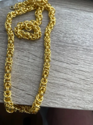 Guldigt kedjehalsband med flätad design - Säljer ett halsband i guldton med en cool flätad kedjedesign. Halsbandet har en chunky look och är tillverkat av metall med glänsande finish. Perfekt för dig som vill ha ett statement-smycke som sticker ut.