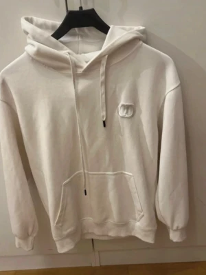 Vit hoodie med broderad logga - Snygg vit hoodie med huva och dragsnören. Har en broderad logga på bröstet och en stor magficka framtill. Tillverkad i mjukt material, perfekt för chill dagar. Ribbade muddar vid ärmslut och nederkant för extra komfort. den är strl s inga skador eller fläckar tar byten beror på vad pris kan alltid diskuteras 