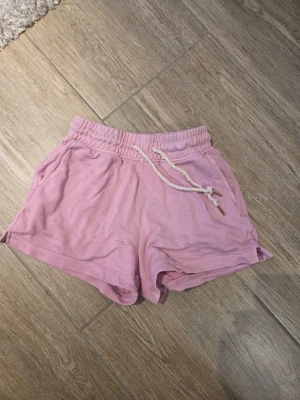 Ljusrosa shorts med snörning - Säljer ett par ljusrosa shorts med elastisk midja och vit snörning i midjan. Shortsen har fickor på sidorna och är tillverkade i mjukt bomullsmaterial. Perfekta för varma dagar eller chill hemma.