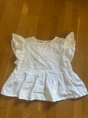 Vit blus med volang och spetsdetaljer - Söt vit blus med broderade spetsdetaljer och volang vid ärmarna. Blusen har en luftig passform och är tillverkad i bomull, perfekt för varma dagar. Blommönstrad brodyr ger en fin känsla, volangärmarna gör toppen extra gullig. Använts några gånger då jag var mindre men är fortfarande väldigt bra skick. 