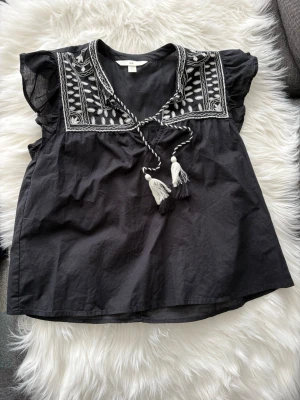Svart broderad blus från H&M - Svart blus från H&M med vita broderier och snörning med tofsar vid halsen. Blusen har korta ärmar med lätt volang och ett boho-inspirerat mönster upptill. Tillverkad i mjuk bomull.