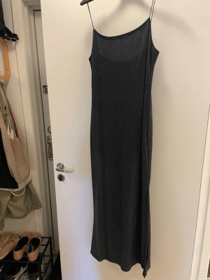 Svart glittrig långklänning Hennes - Svart långklänning med tunna axelband från Hennes. Klänningen har en glittrig finish och en smal, rak passform som ger en elegant look. Perfekt för dig som vill sticka ut med stilrena detaljer och enkel design.