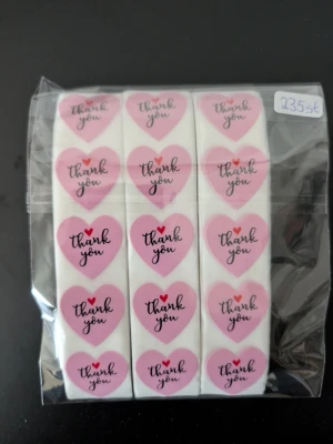 Hjärtformade thank you-klistermärken - Ett paket med 235 stycken hjärtformade klistermärken i rosa och vitt med texten 'thank you' och en liten röd hjärtdetalj. Perfekta för paket, brev eller presenter när du vill säga tack på ett gulligt sätt.