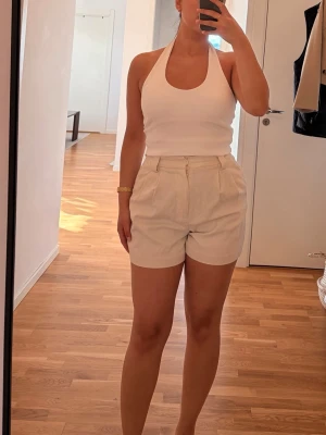 Beige högmidjade linneshorts - Endast tvättade och testade, ej använda. Snygga beige shorts i linneliknande material med hög midja och avslappnad passform. De har dragkedja framtill, bälteshällor och två sidofickor. Perfekta för varma dagar och ger en clean och fräsch look.