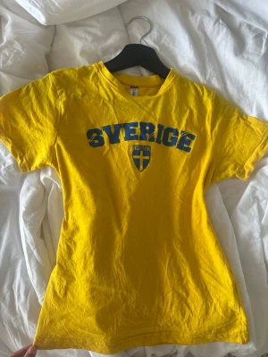Gul Sverige t-shirt från Clique - Gul t-shirt från Clique med blå texten 'Sverige' och svensk flagga på bröstet. Klassisk passform och rund halsringning. Tillverkad i mjuk bomull så den är så skön! Strlk 130/140 men sitter som en XXS/XS