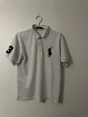 Vit pikétröja med mörkblå logga - Klassisk vit pikétröja med krage och knappar framtill. Stor mörkblå Ralph Lauren logga på bröstet och siffran 3 på ena ärmen. Kortärmad och i ett mjukt bomullsmaterial som passar perfekt till jeans eller shorts.