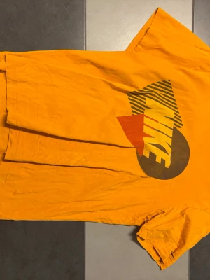 Gul Nike t-shirt med retrotryck - Gul t-shirt från Nike med stort retroinspirerat tryck i svart, rött och gult på ryggen. Klassisk passform och korta ärmar. Perfekt för dig som gillar färgstarka plagg och streetwear-vibbar.