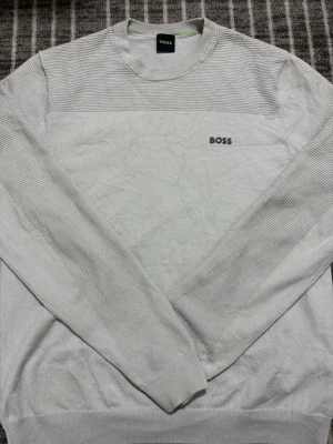 Ljusgrå tröja från BOSS - Snygg ljusgrå tröja från BOSS med diskret logga på bröstet. Tröjan har rund halsringning, ribbade partier på axlar och ärmar samt en stilren och minimalistisk design. Perfekt för dig som gillar klassisk och enkel stil. Pris kan diskuteras vid snabb affär