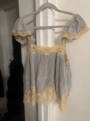 Ljusgrå offshoulder linnetopp med spets - Supersöt ljusgrå linnetopp med offshoulder-ärmar och beige spetsdetaljer längs kanterna. Toppen har en lös passform och är tillverkad i ett tunt, silkesliknande material som ger en romantisk vibe. Perfekt för sommarens alla tillfällen.