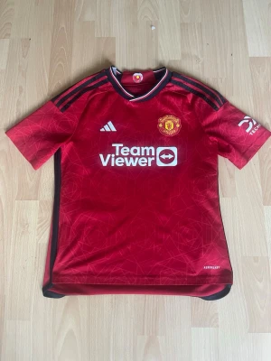 Manchester United tröja aitentisk - Säljer en röd Manchester United tröja från Adidas med svarta detaljer och klubbmärke på bröstet. Tröjan har korta ärmar, rund hals. Tillverkad i lätt och ventilerande material, perfekt för fotboll eller träning. Jag säljer eftersom att den är för liten. Den är i storlek 164cm