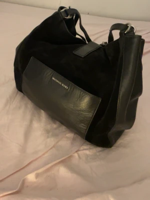 Svart handväska från Michael Kors - Stilren svart handväska från Michael Kors i en kombination av mocka och slätt läder. Väskan har silverfärgade metalldetaljer, en stor ytterficka framtill med logga och rundade former. Perfekt för dig som gillar klassisk och tidlös design.
