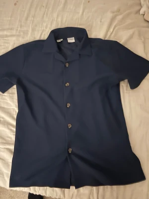 Mörkblå kortärmad skjorta med knappar - Snygg mörkblå skjorta i polyester med korta ärmar och klassisk krage. Skjortan har knappar framtill och en diskret ribbad struktur i tyget som ger en modern känsla. Perfekt för dig som vill ha en enkel men stilren look.