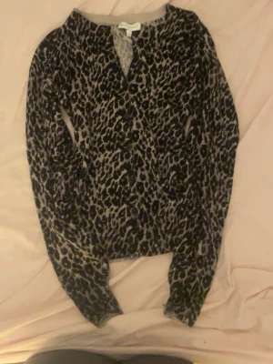 Leopardmönstrad kofta från Warehouse - Snygg leopardmönstrad kofta från Warehouse med knappar framtill. Koftan har långa ärmar och ribbad kant vid halsen. Färgerna går i svart och beige, vilket ger en trendig och djurinspirerad look.