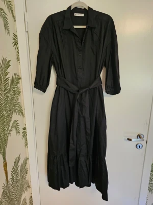 Midiklänning med knytskärp - Svart midiklänning modell Vendela i oversized snitt med krage, knappar framtill och trekvartsärm. Klänningen har ett matchande knytskärp i midjan och volang nedtill. Kan bäras både med eller utan skärp. 