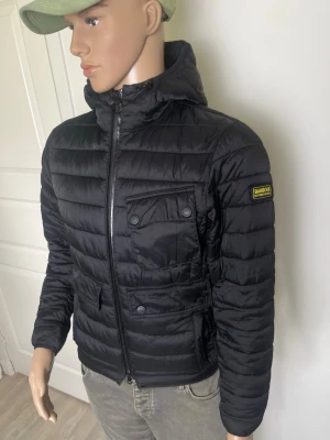 Svart dunjacka från Barbour storlek s - Snygg svart dunjacka från Barbour International med quiltad design, huva och två stora bröstfickor med knappar. Jackan har dragkedja framtill och Barbour-logga på ärmen. Perfekt för kalla vinterdagar och riktigt stilren look.