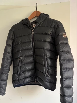 Svart dunjacka från Colmar - Snygg svart dunjacka från Colmar med huva och dragkedja framtill. Jackan har två snedställda fickor med dragkedja och Colmar-logga på ärmen. Säljer då den inte kommer t användning och är lite liten för mig, skriv vid intresse