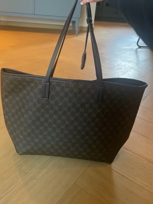 Mörkbrun shoppingväska Don Donna - Snygg mörkbrun shoppingväska från Don Donna med monogrammönster över hela väskan. Tillverkad i konstläder med detaljer i samma färg. OBS: se sista bild för defekt på ena handtaget!! Däremot ej så synligt och påverkar inte användningen. Köpt för 499kr🫶🏻rymmer dator, kan skicka mått vid förfrågning!! 
