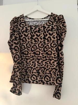 Leopardmönstrad långärmad topp - Säljer en snygg topp med leopardmönster i beige och svart. Toppen har puffiga axlar och långa ärmar som ger en trendig look. Materialet känns mjukt och stretchigt, perfekt för dig som vill sticka ut lite extra. Aldrig använd strl L