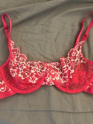Rosa spets-bh med blommönster - Säljer en snygg rosa bh med vita och rosa blommiga spetsdetaljer. Bh:n har justerbara axelband och bygel för extra stöd. Materialet är genomskinlig spets som ger en feminin och trendig look. Perfekt för dig som gillar fina detaljer och vill ha något extra i underklädeslådan.